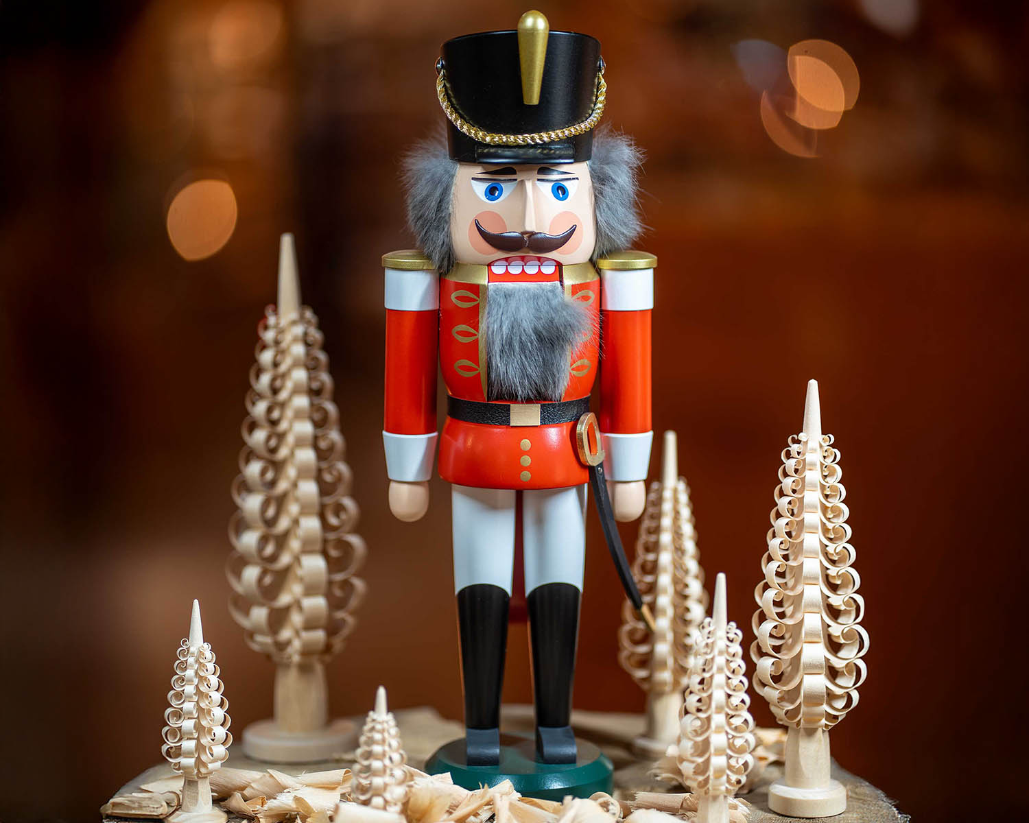 Ein klassischer erzgebirgischer Nussknacker aus Holz in roter Uniform mit goldenen Verzierungen, schwarzem Hut mit goldener Kordel, grauem Bart und blauen Augen. Er steht vor mehreren filigran gedrechselten Spanbäumchen in verschiedenen Größen. Im Hintergrund warmes, unscharfes Licht und Holzspäne am Fuß