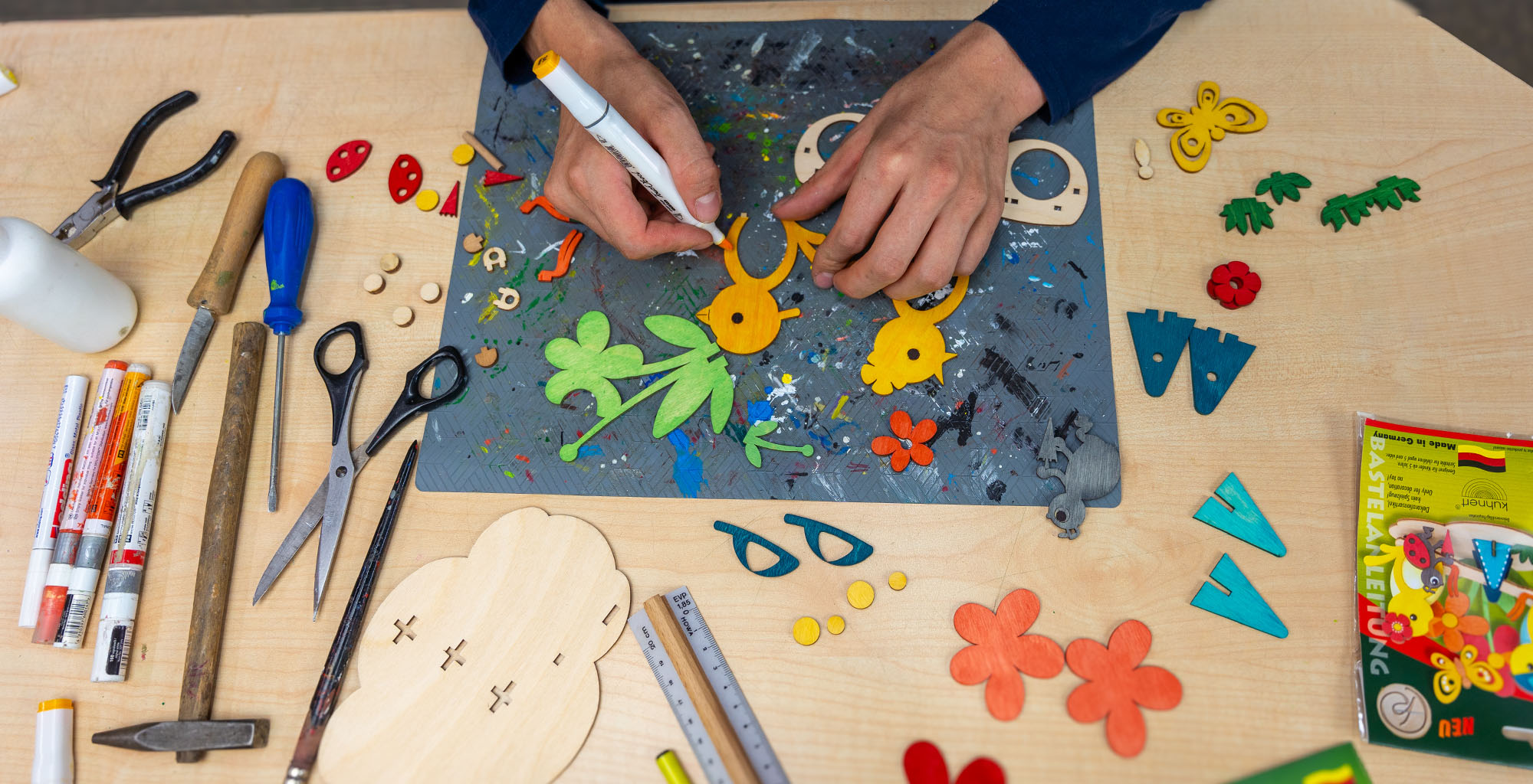 Zwei Hände basteln an einem Kunstwerk mit gelben Vögeln grünen Blättern und bunten Blumen auf einer bemalten Unterlage Werkzeuge wie Schere Zangen und Marker liegen daneben