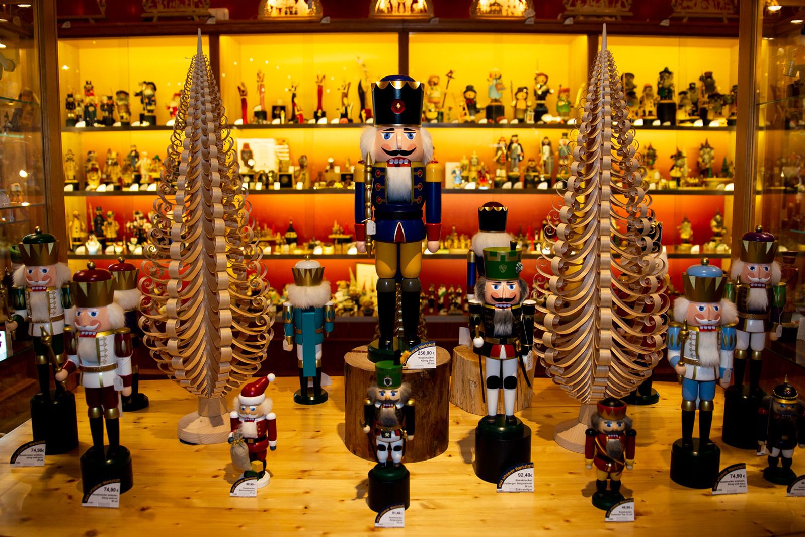 Ein Raum mit Holzregalen voller Nussknackerfiguren in verschiedenen Größen und Farben Vorne stehen große Nussknacker mit Uniformen zwischen dekorativen Tannenbäumen aus Holz Hintergrund beleuchtet mit orangem Licht