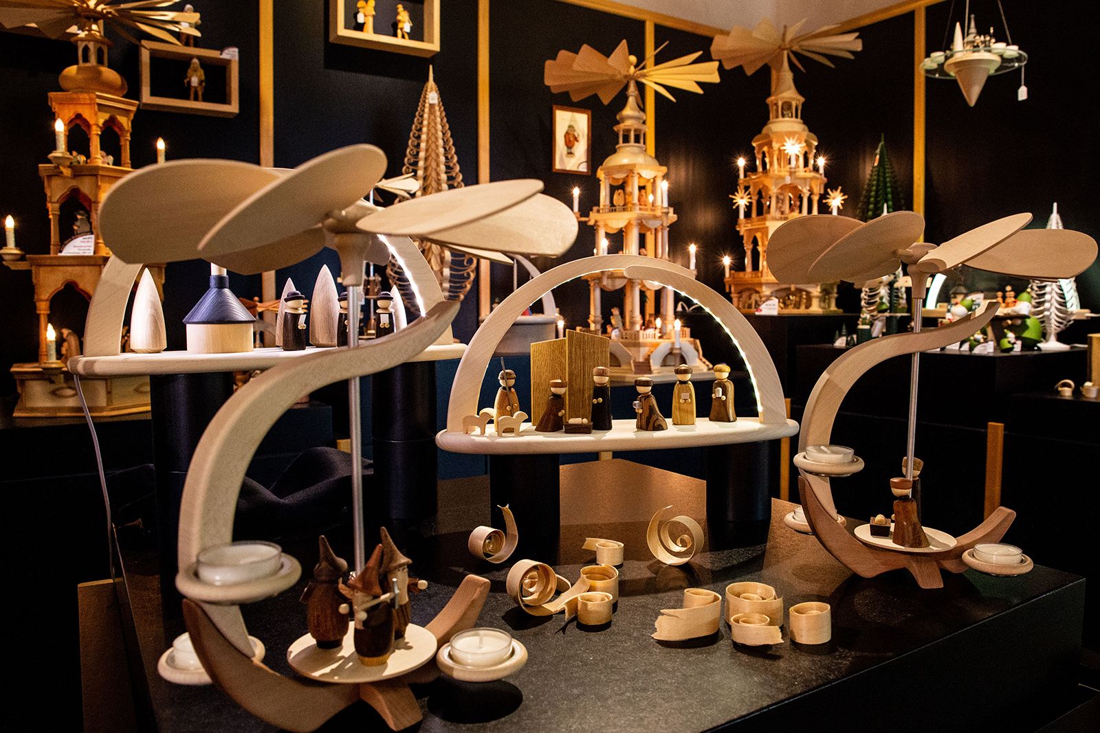 Ein Raum mit mehreren Weihnachtspyramiden aus Holz mit rotierenden Flügeln Darauf Figuren und Kerzenhalter Dekorationen wie Tannenbäume und Lichter dunkler Hintergrund