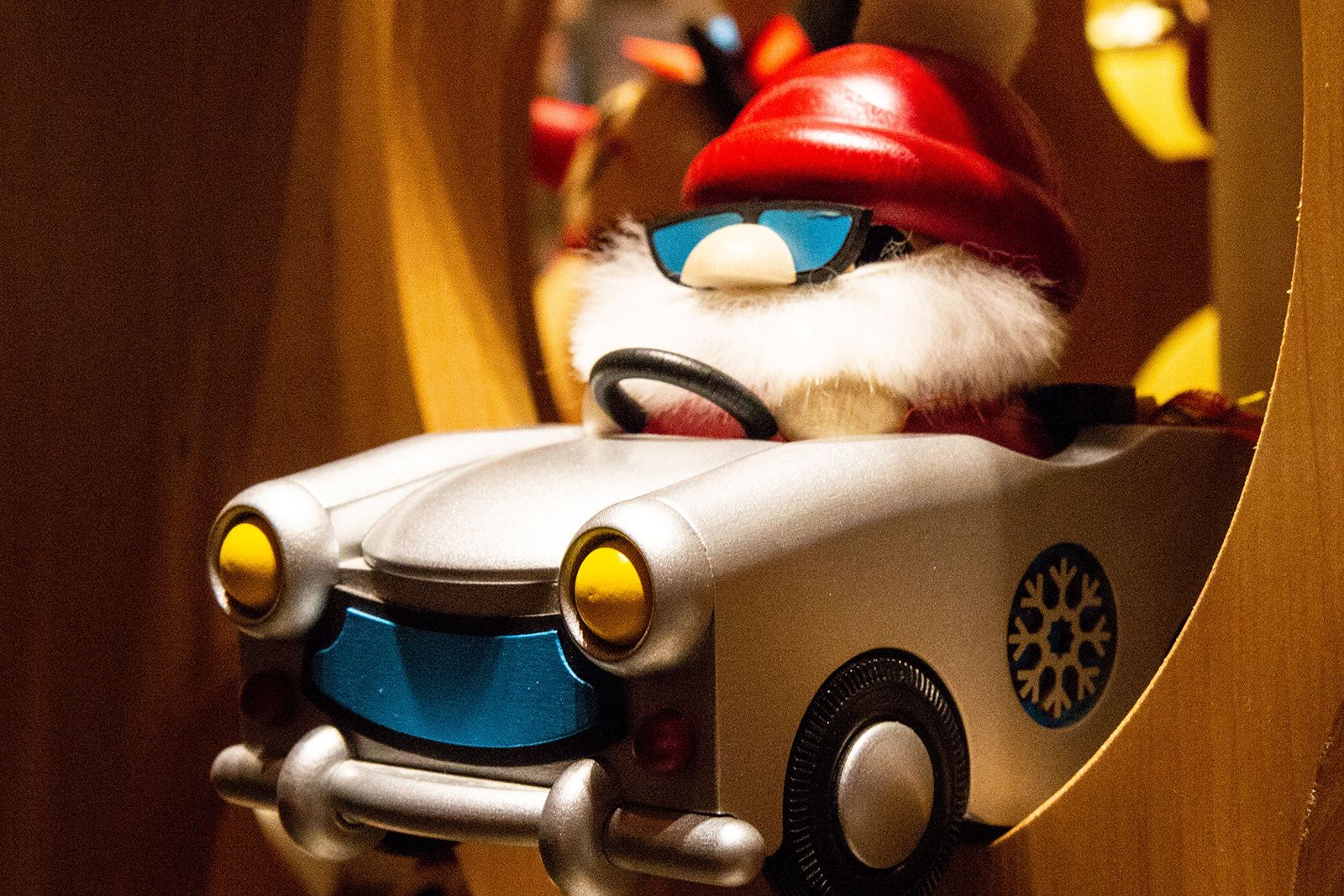 Eine Räucherfigur mit rotem Hut und weißem Bart sitzt in einem silbernen Trabi Figur trägt Sonnenbrille Auto hat gelbe Scheinwerfer Hintergrund holzfarben