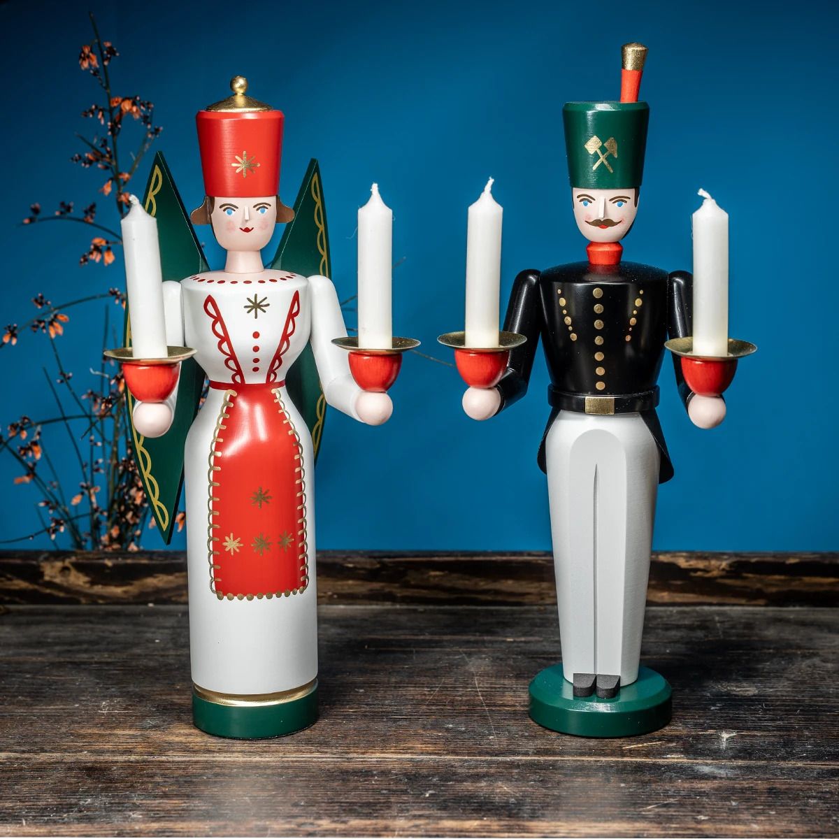 Zwei hölzerne Figuren mit Kerzen ein Engel in rot-weiß und ein Bergmann in schwarz-weiß mit grünem Hut stehen auf einer Holzfläche Im Hintergrund blauer Untergrund und ein Ast mit Blüten