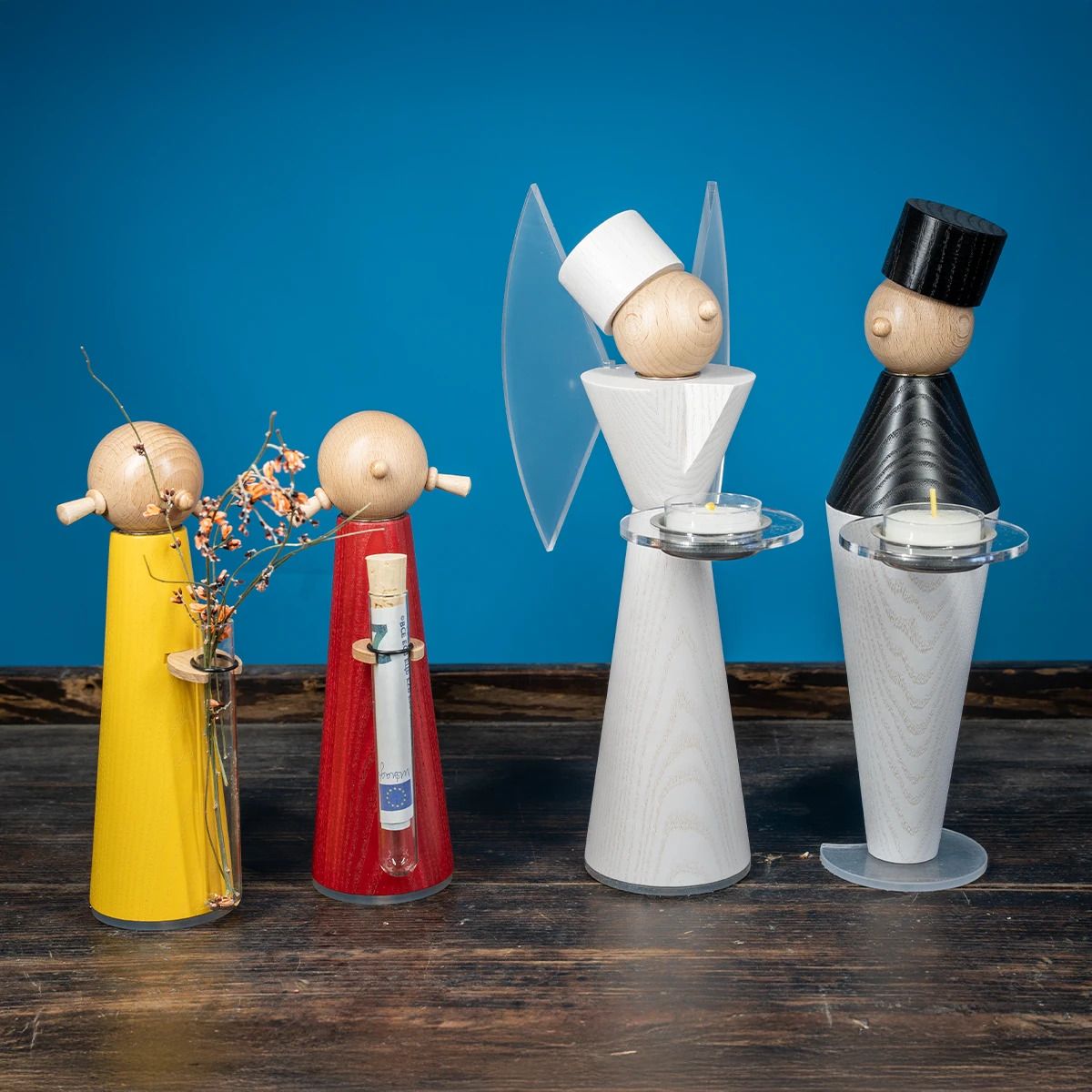 Vier moderne hölzerne Figuren ohne Gesicht stehen auf einer Holzfläche Zwei Mädchen in gelb und rot mit Vasen und trockenen Blüten dazu ein Engel und Bergmann weiß und schwarz mit Kerzenhaltern Im Hintergrund blauer Untergrund