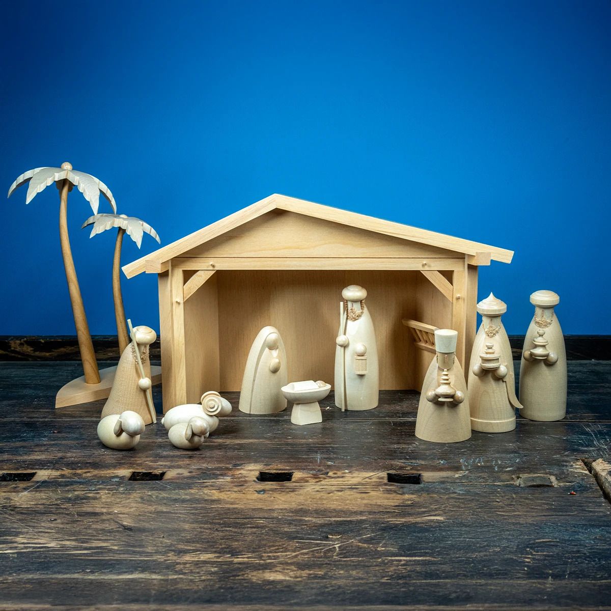 Eine hölzerne Krippe mit Figuren steht auf einer Holzfläche Dazu gehören sechs Personen ein Krippenstall zwei Schafe und zwei Palmen Im Hintergrund blauer Untergrund