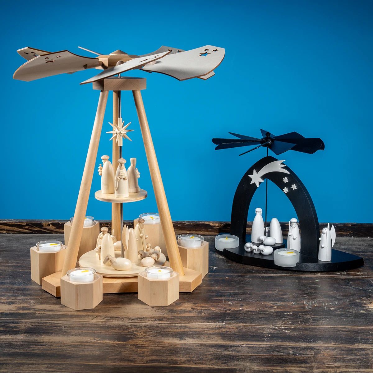 Eine hölzerne Pyramide mit Flügeln und Figuren sowie Kerzenhaltern steht auf einer Holzfläche Daneben eine schwarze Pyramide mit weißen Figuren und Sternen Im Hintergrund blauer Untergrund