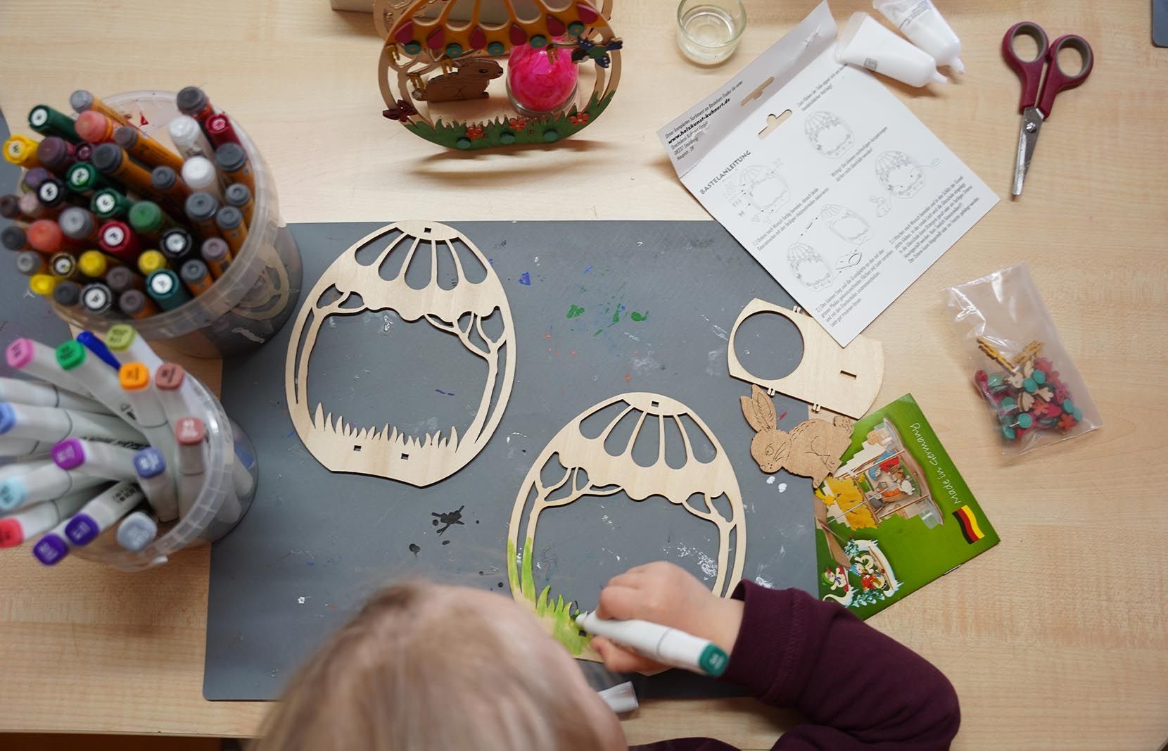 Ein Kind malt mit einem grünen Stift an einem Holzmodell eines Ostereis mit Ornamenten. Auf dem Tisch liegt ein weiteres unbemaltes Ei-Modell, Farbstifte, Kleber, Schere und eine Anleitung. Im Hintergrund steht ein fertiges Modell.