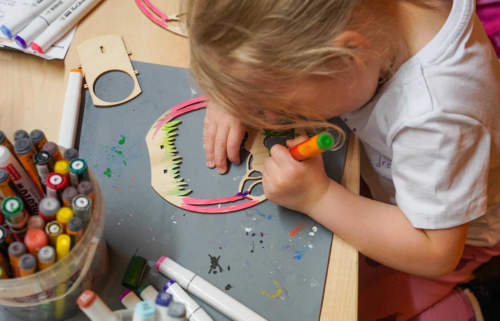 Ein Kind in weißem Shirt malt mit einem orangefarbenen Stift an einem Holzmodell eines Ostereis mit Ornamenten. Auf dem Tisch stehen Farbstifte. Farbspritzer sind auf der Unterlage sichtbar.