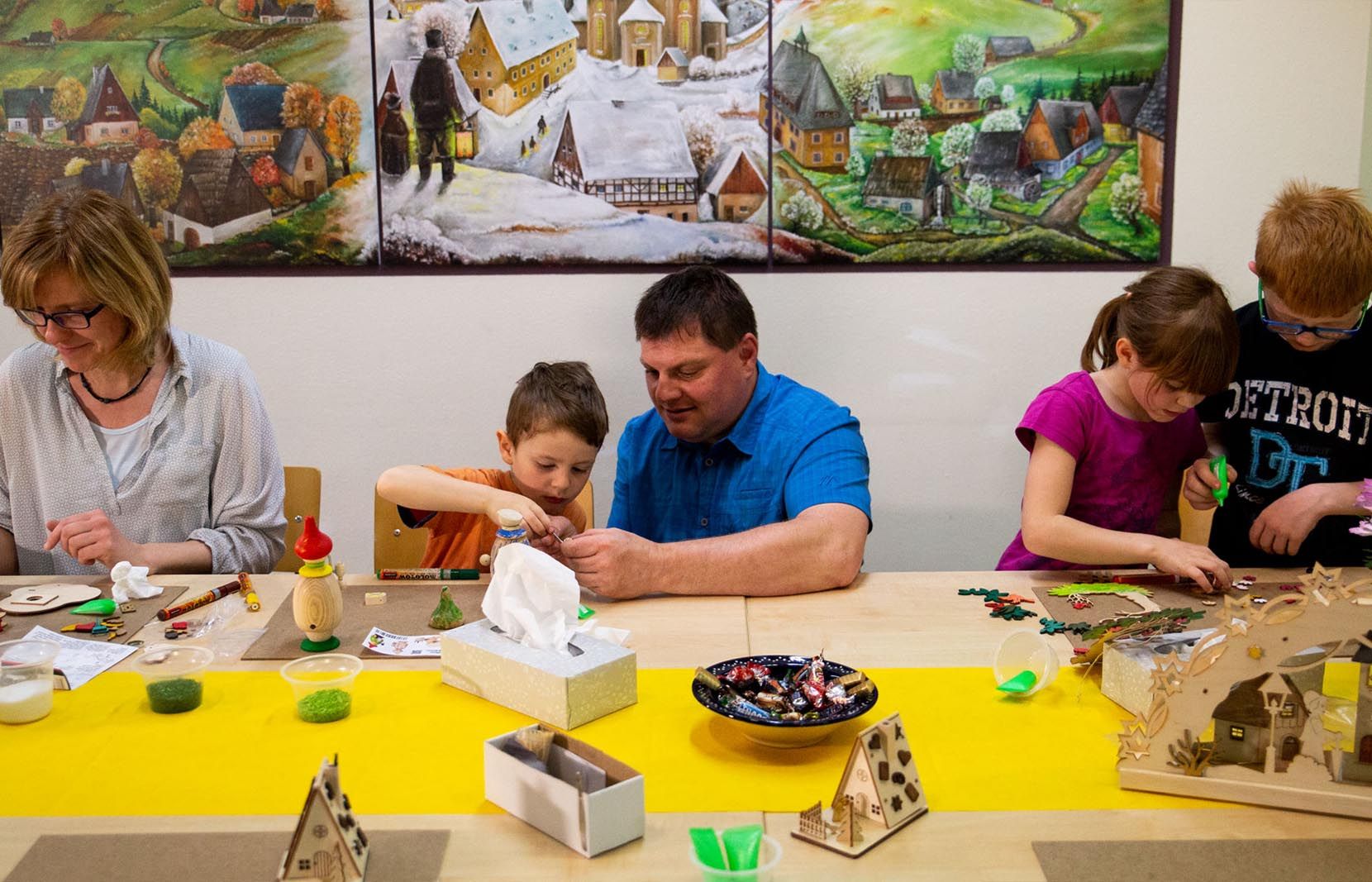Zwei Erwachsene und drei Kinder sitzen an einem Tisch und basteln an Holzmodellen, darunter Häuser. Auf dem Tisch stehen verschiedene Bastelmaterialien. Im Hintergrund hängt ein Bild mit erzgebirgischer Landschaft.