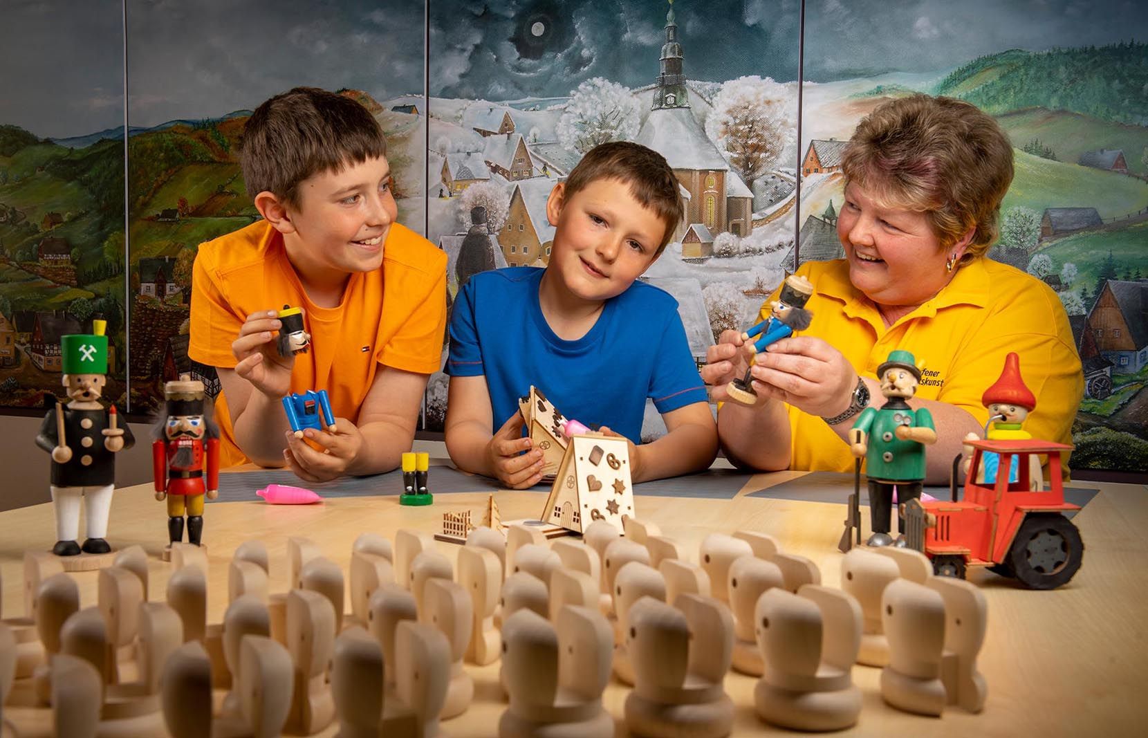 Drei Personen, zwei Kinder in orange und blauem Shirt und eine Erwachsene in gelbem Shirt, sitzen an einem Tisch und halten Holzfiguren wie Nussknacker und Räuchermännchen. Vor ihnen liegen unbemalte Holzstücke und ein kleines Hausmodell. Im Hintergrund ein Gemälde mit winterlicher Landschaft.