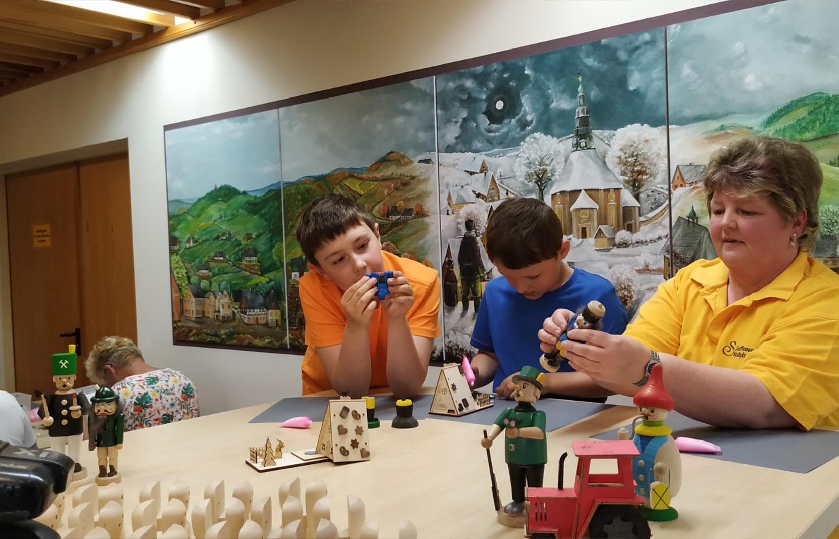 Drei Kinder und eine Erwachsene in gelbem Shirt sitzen an einem Tisch und halten Holzfiguren wie Nussknacker und Räuchermännchen. Vor ihnen stehen kleine Hausmodelle und ein roter Traktor. Im Hintergrund ein Gemälde mit winterlicher Landschaft und Seiffener Dorf.