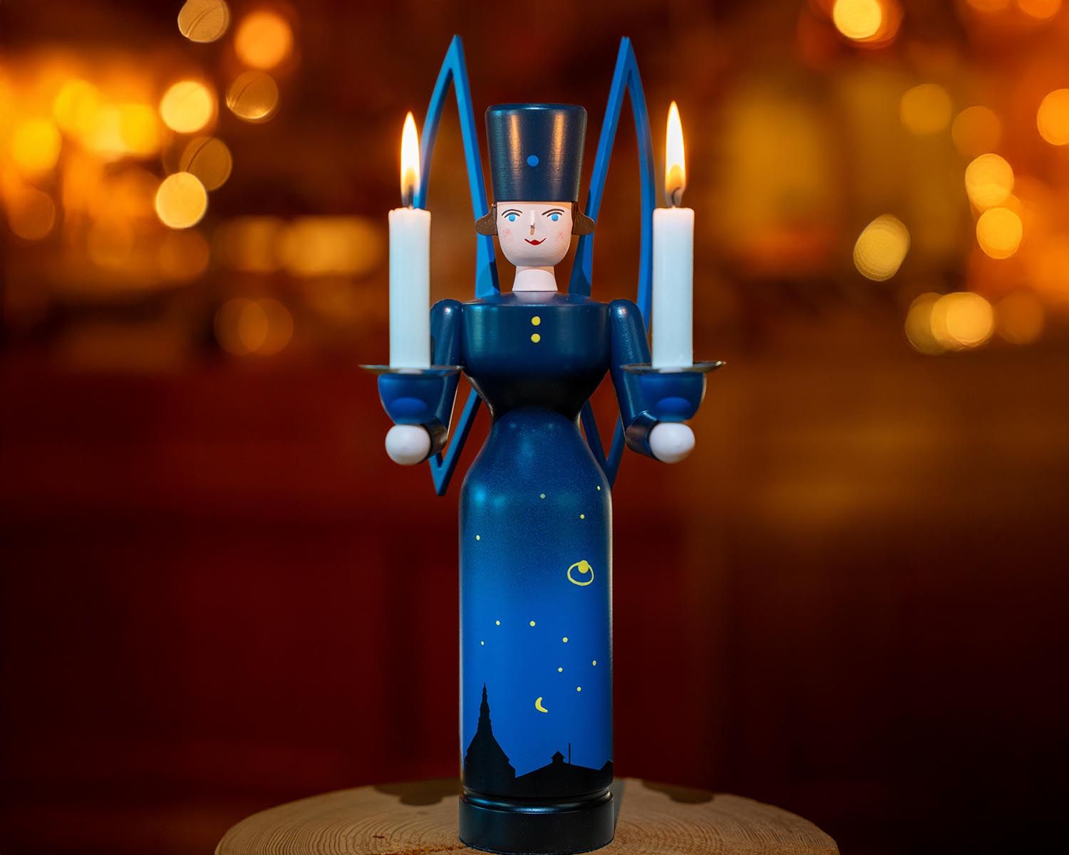 Ein blauer erzgebirgischer Engel aus Holz mit schwarzem Hut und goldenem Knopf, der zwei brennende weiße Kerzen in den Armen hält. Der Körper ist mit einem Nachthimmel-Motiv bemalt: goldene Sterne, Mondsichel und die Silhouette der Seiffener Kirche. Im Hintergrund unscharfes, warmes Licht