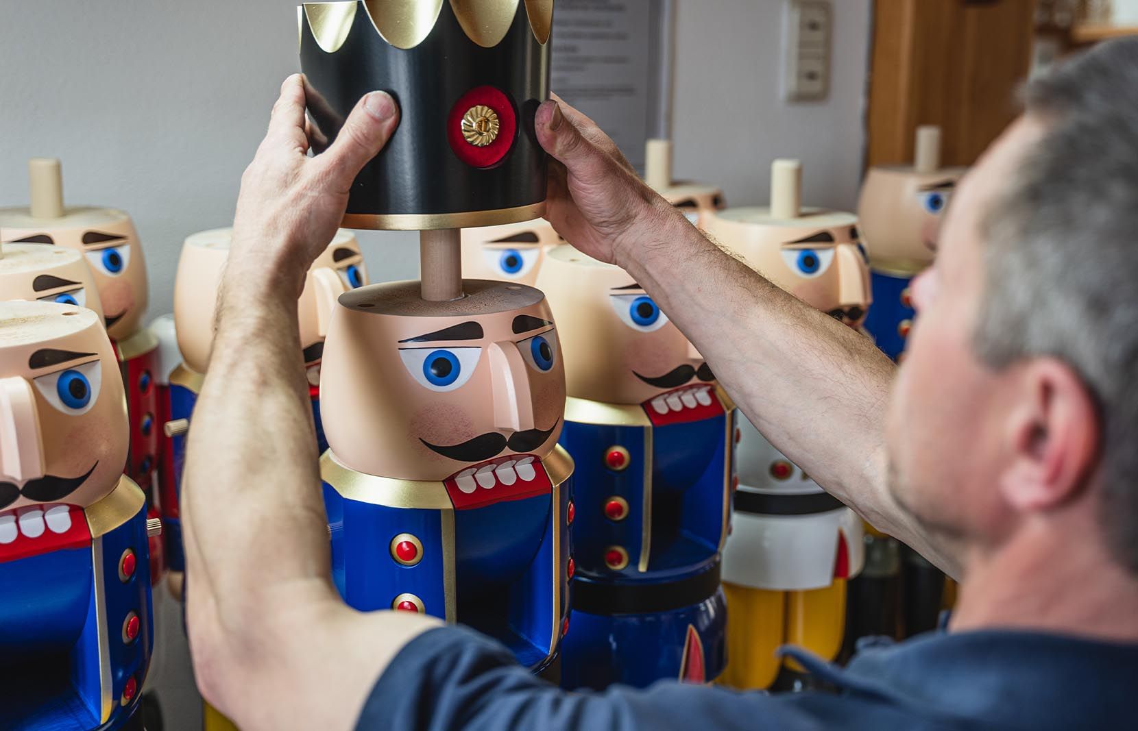 Eine Person mit dunkelblauer Kleidung setzt einem bunt bemalten Nussknacker König eine Krone auf welche goldene Verzierungen hat