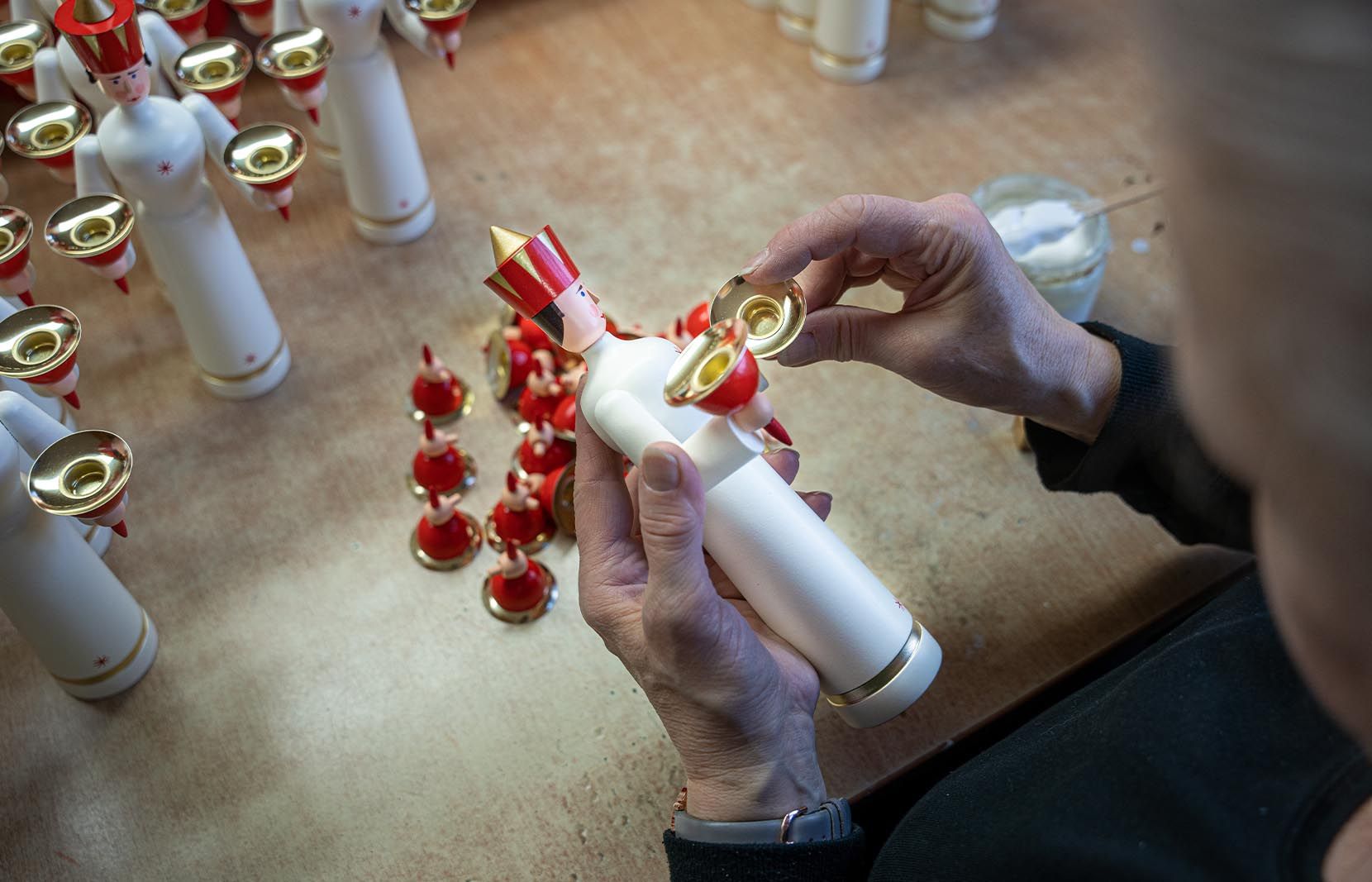 Eine Hand montiert eine weiße Engelsfigur mit rotem Hut und goldenen Kerzenhaltern. Auf dem Tisch stehen weitere Engel und rote Deko-Elemente.