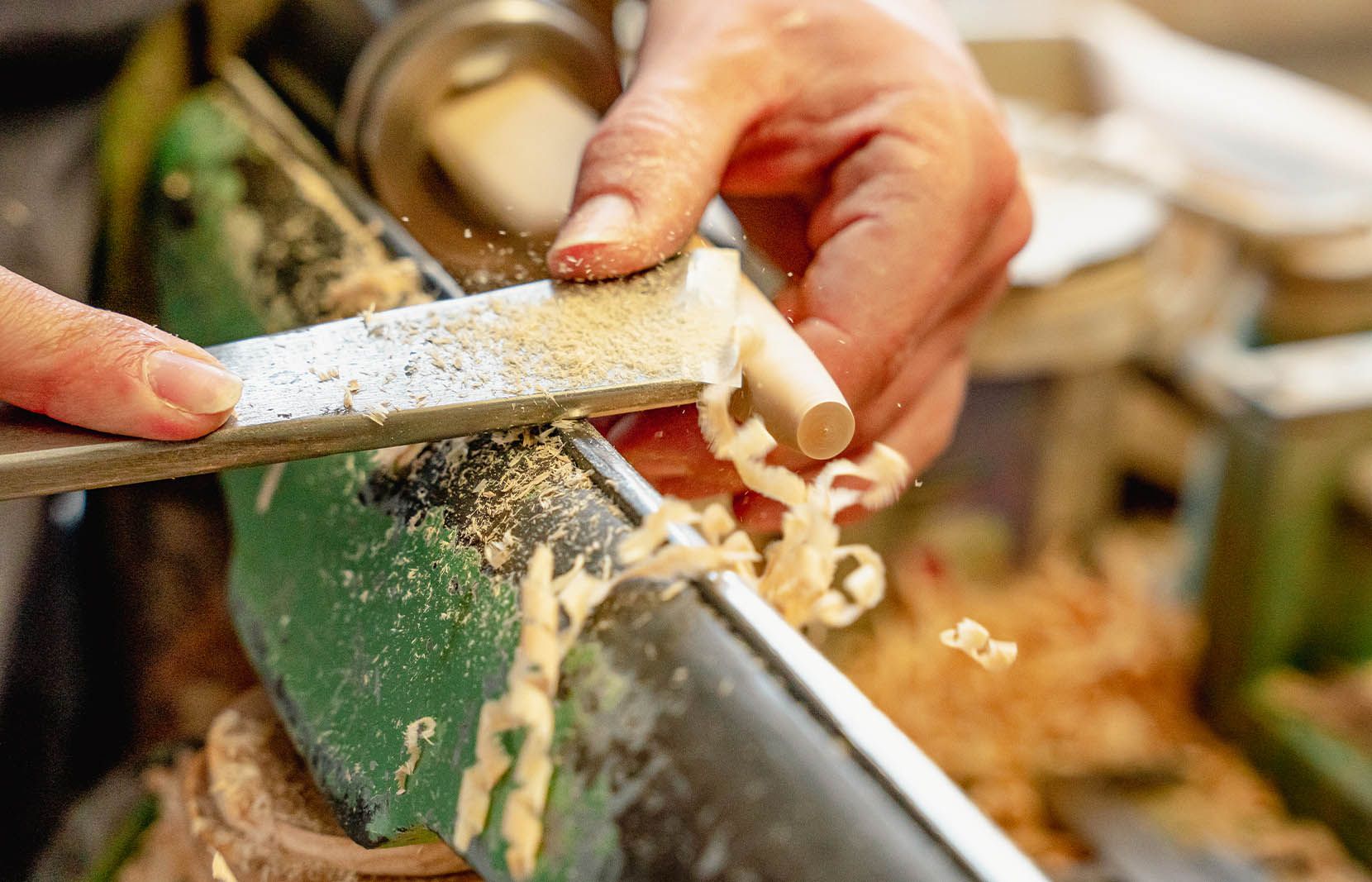 Zwei Hände bearbeiten ein Holzstück an einer grünen Drehbank. Mit einem Meißel wird das Holz geformt, während Holzspäne abfallen.