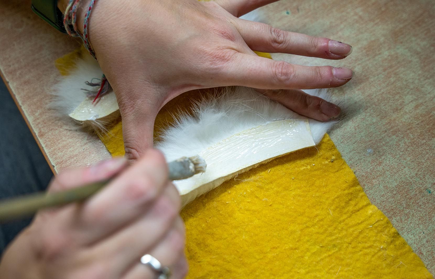 Eine Hand bestreicht mit einem Pinsel einen weißen Bart aus Fell mit Leim. Die Unterlage ist gelb. Der Vorgang findet auf einer holzfarbenen Oberfläche statt.