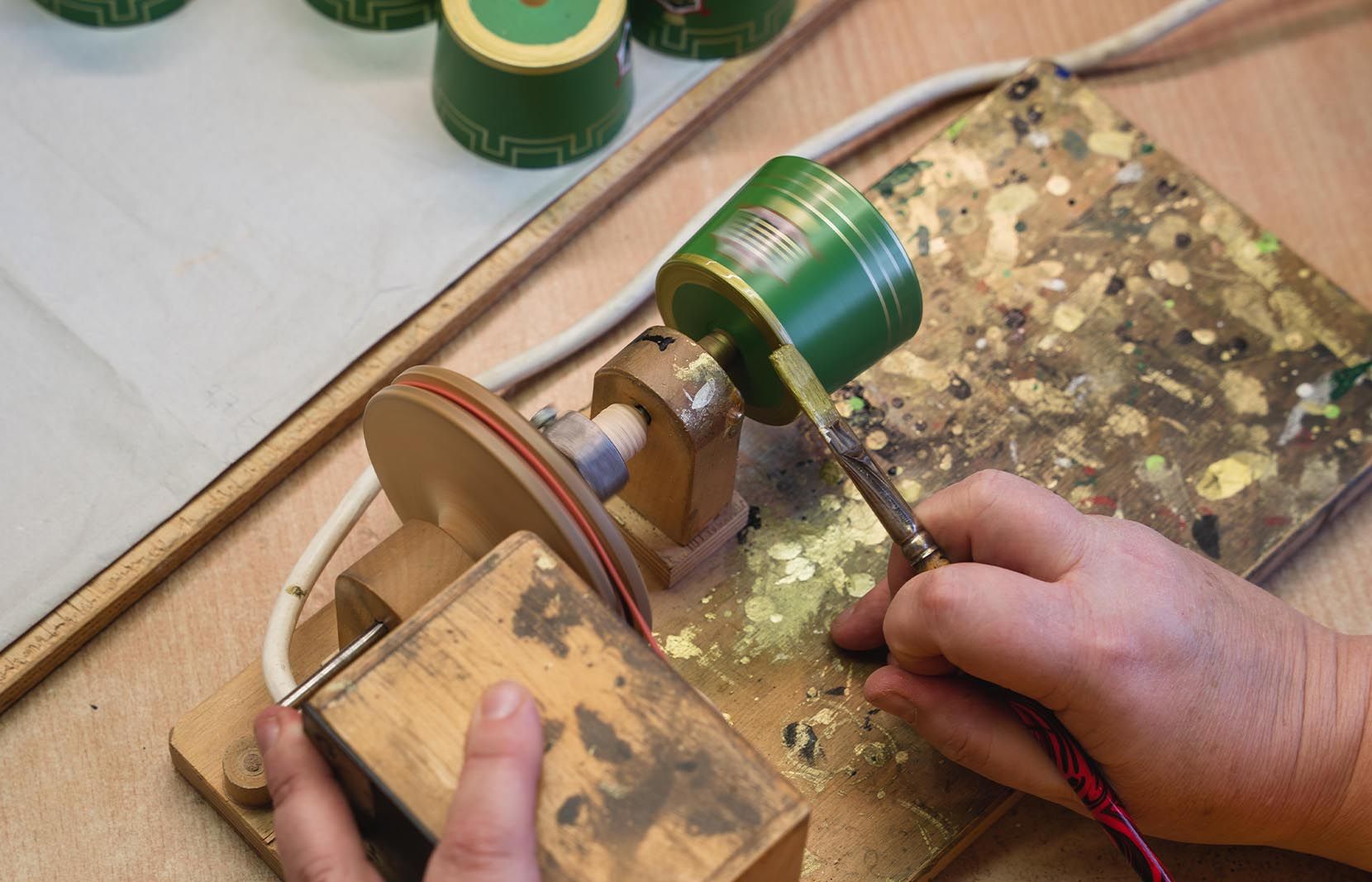 Zwei Hände arbeiten an einem grünen Holz-Hut auf einer Drehbank. Eine Hand hält die Drehbank, die andere führt einen Pinsel. Im Hintergrund sind weitere grüne Hüte sichtbar. Die Arbeitsfläche ist holzfarben und mit Farbresten bedeckt.