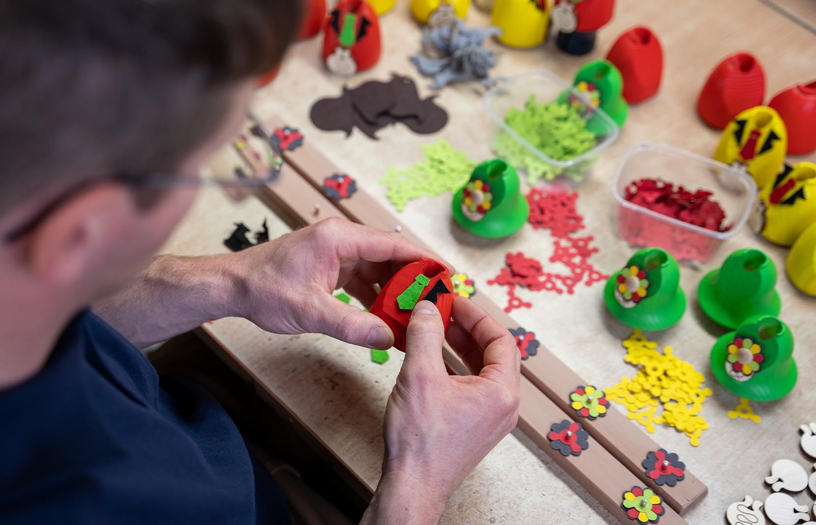 Eine Person in dunkelblauem Hemd montiert bunte Holzteile auf einem Tisch mit roten grünen und gelben Figuren und Zubehörteilen
