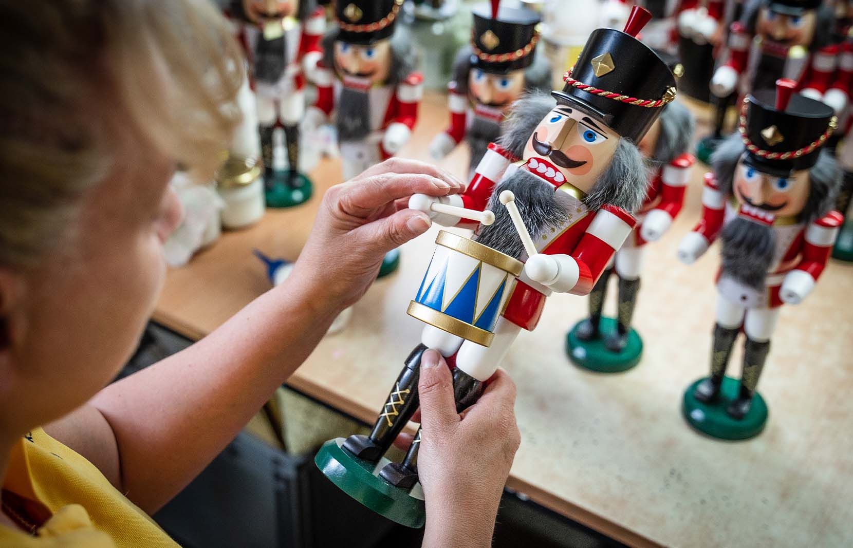 Eine Person hält einen bunt bemalten Nussknacker welcher graues Haar hat und eine Trommel in der Hand hält im Hintergrund eine Reihe ähnlicher Nussknacker verschwommen sichtbar