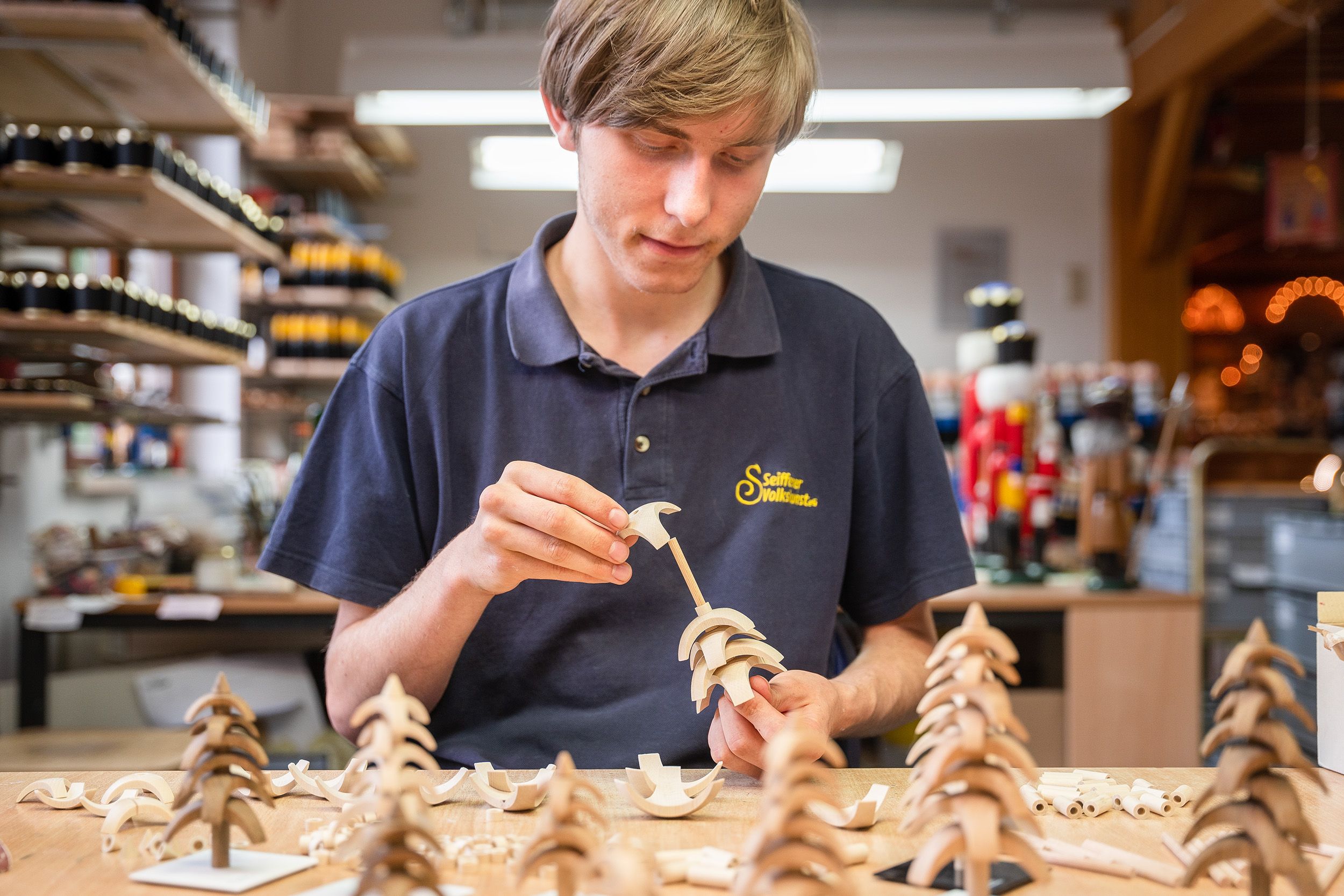 Ein junger Mann in einem blauen Poloshirt mit "Seiffener Volkskunst" Logo arbeitet an einem modernen Holzbaum. Er montiert Holzstücke an einem Stab. Auf dem Tisch liegen mehrere fertige und unfertige Holzbäume sowie lose Holzstücke. Im Hintergrund sind Regale mit Materialien und weitere Holzfiguren sichtbar.