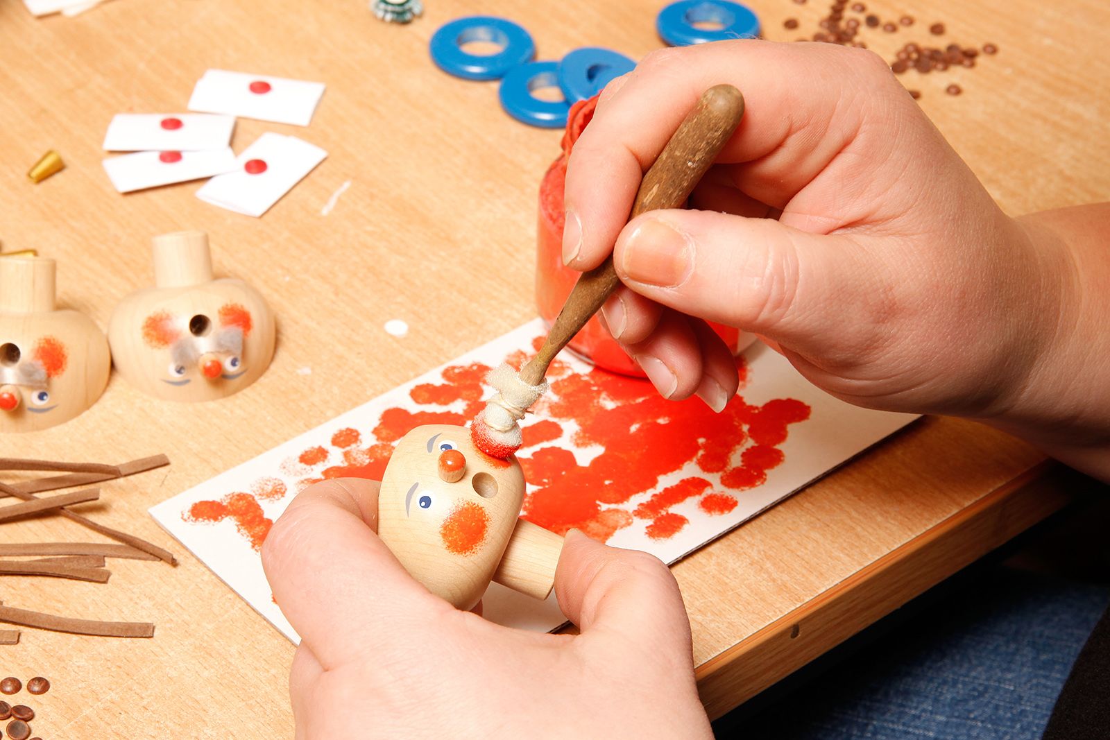 Zwei Hände bemalen die Wangen einer Holzfigur mit rotem Pinsel auf einem Tisch mit Werkzeugen kleinen Teilen und blauen Ringen