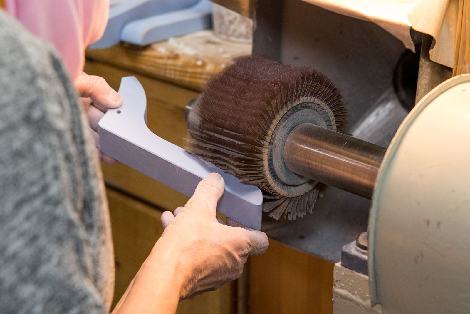 Zwei Hände halten ein Werkstück an eine rotierende Bürste einer Schleifmaschine Holzregal und graue Kleidung sichtbar