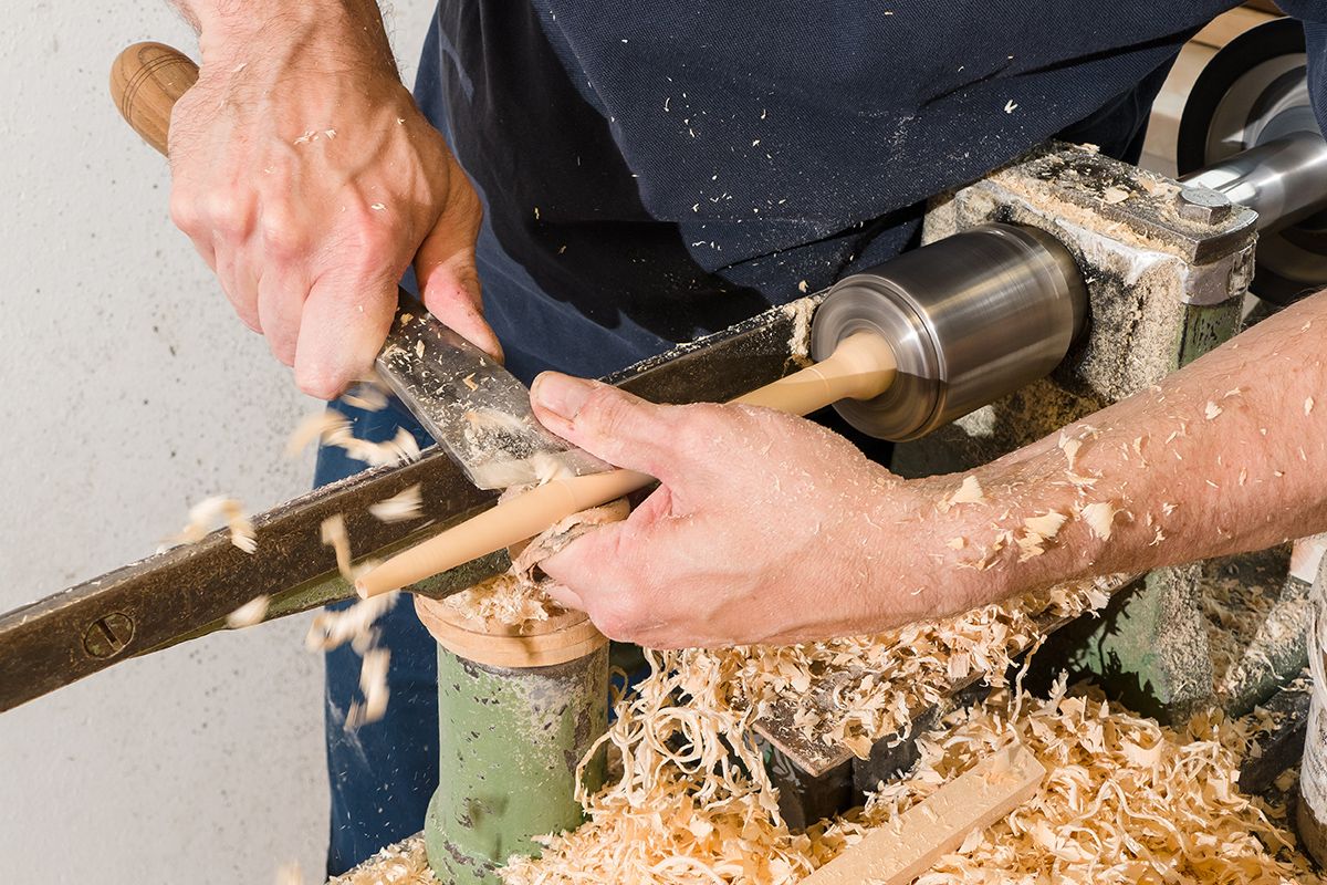 Zwei Hände bearbeiten Holz an einer Drehbank mit Werkzeug Holzspäne fliegen herum grüner Metallrahmen dunkle Kleidung sichtbar