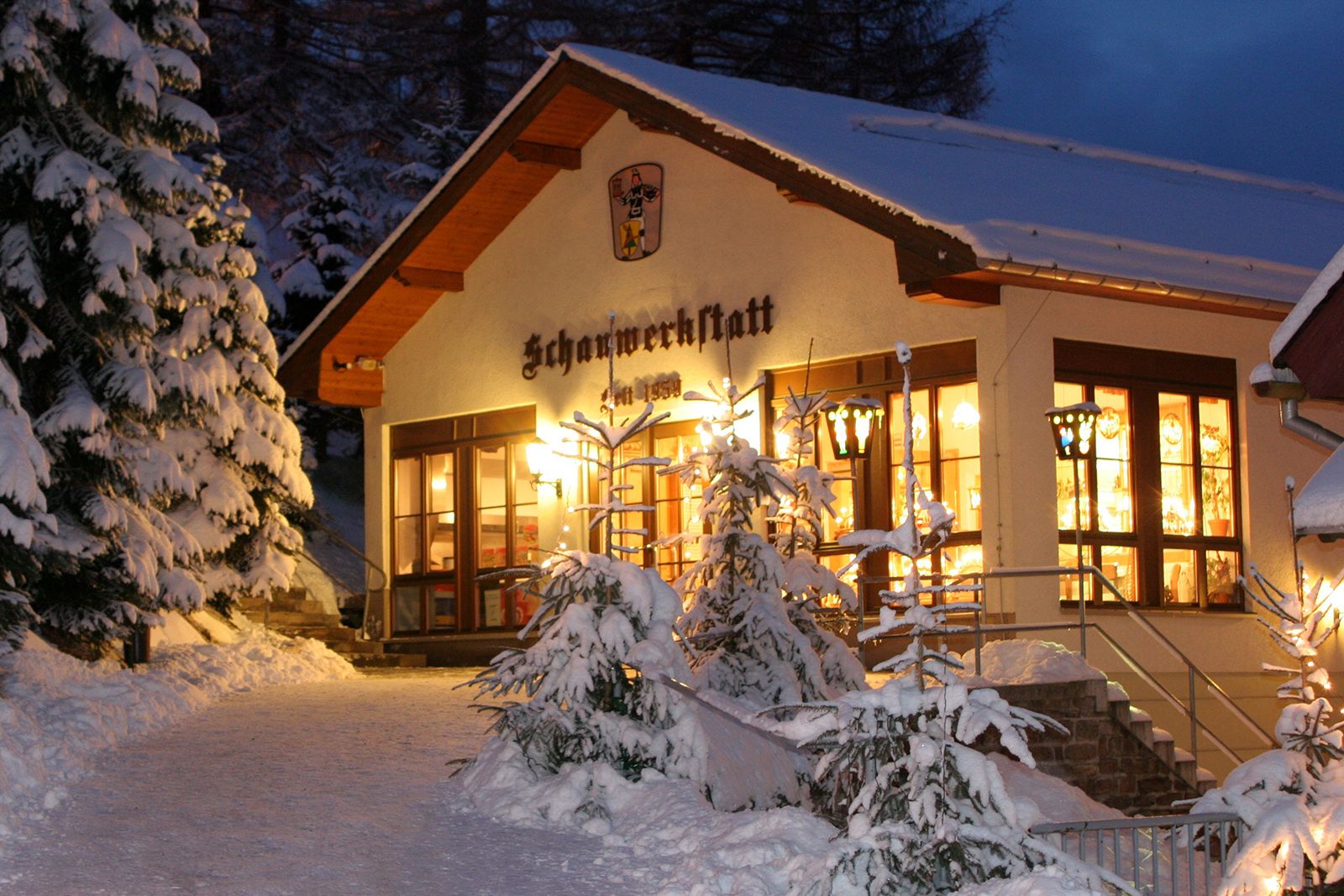 Ein beleuchtetes Gebäude mit dem Schriftzug "Schauwerkstatt" und dem Jahr "1958" bei Nacht im Schnee. Es hat einen steilen, verschneiten Dachfirst, Holzverkleidung und große Fenster, durch die warmes Licht scheint. Vor dem Gebäude stehen verschneite Tannenbäume und eine Treppe führt hinauf. Ein Wappen mit Bergmannsfigur ziert die Fassade.
