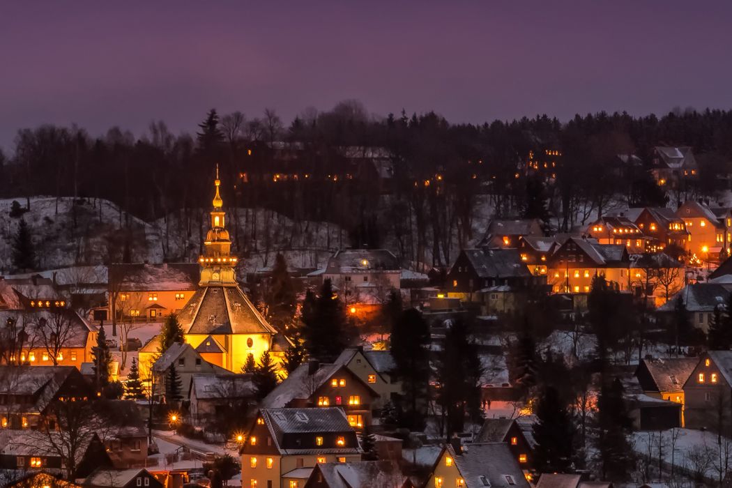 Der verschneite Ort Seiffen bei Nacht mit beleuchteten Häusern und einer runden Kirche Im Hintergrund dunkler Wald und lila Himmel