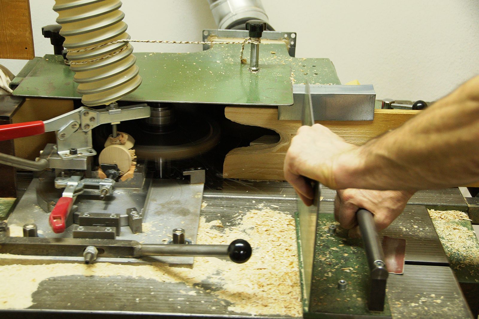 Eine Hand schiebt ein Holzstück durch eine grüne Fräsmaschine mit rotem Hebel gelber Schlauch und Holzspäne sichtbar