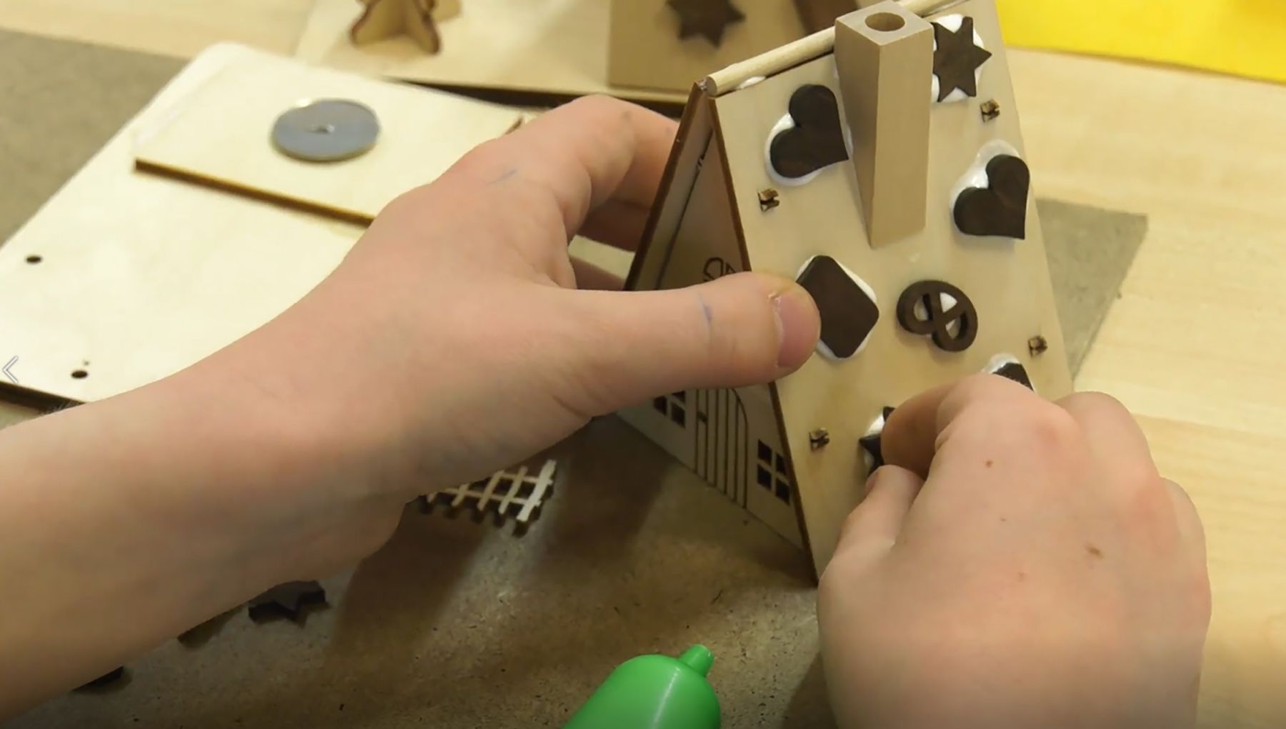 Zwei Hände basteln ein kleines Holzhaus mit Lebkuchen an den Wänden. Eine Hand hält das Haus, die andere verwendet einen Kleber. Auf dem Tisch liegen weitere Holzstücke.