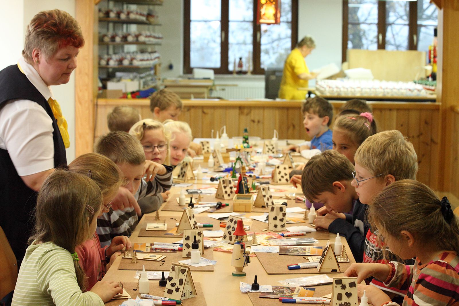 Eine Gruppe Kinder sitzt an einem langen Tisch und bastelt an kleinen Holzmodellen, darunter Häuser und Figuren. Eine Erwachsene in gelb-schwarzer Kleidung steht daneben und überwacht. Der Raum hat Holzverkleidung, Regale mit Materialien und Fenster mit Blick nach draußen.